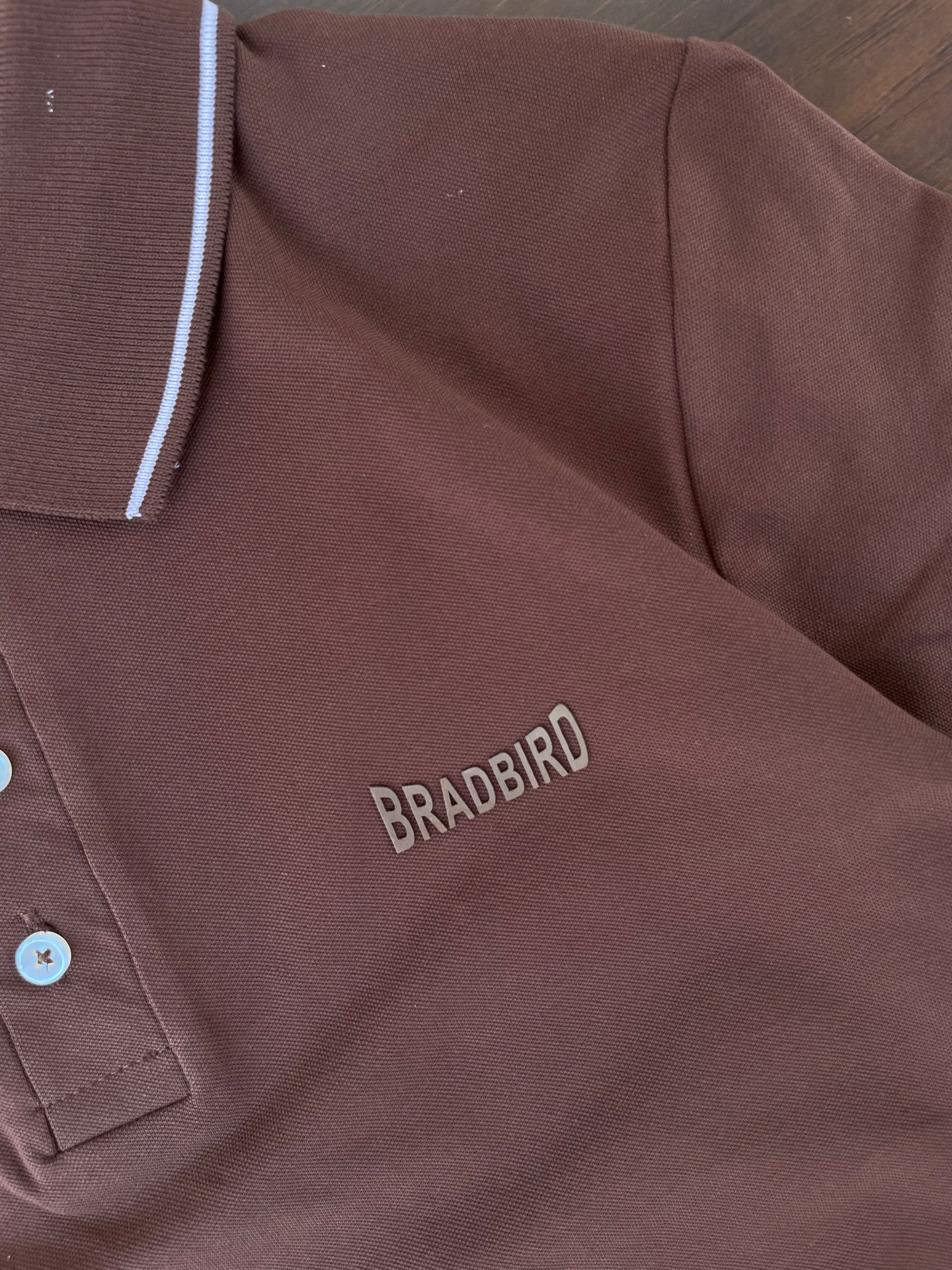 Close-up of brown polo chest with BRADBIRD logo and white-trim buttons