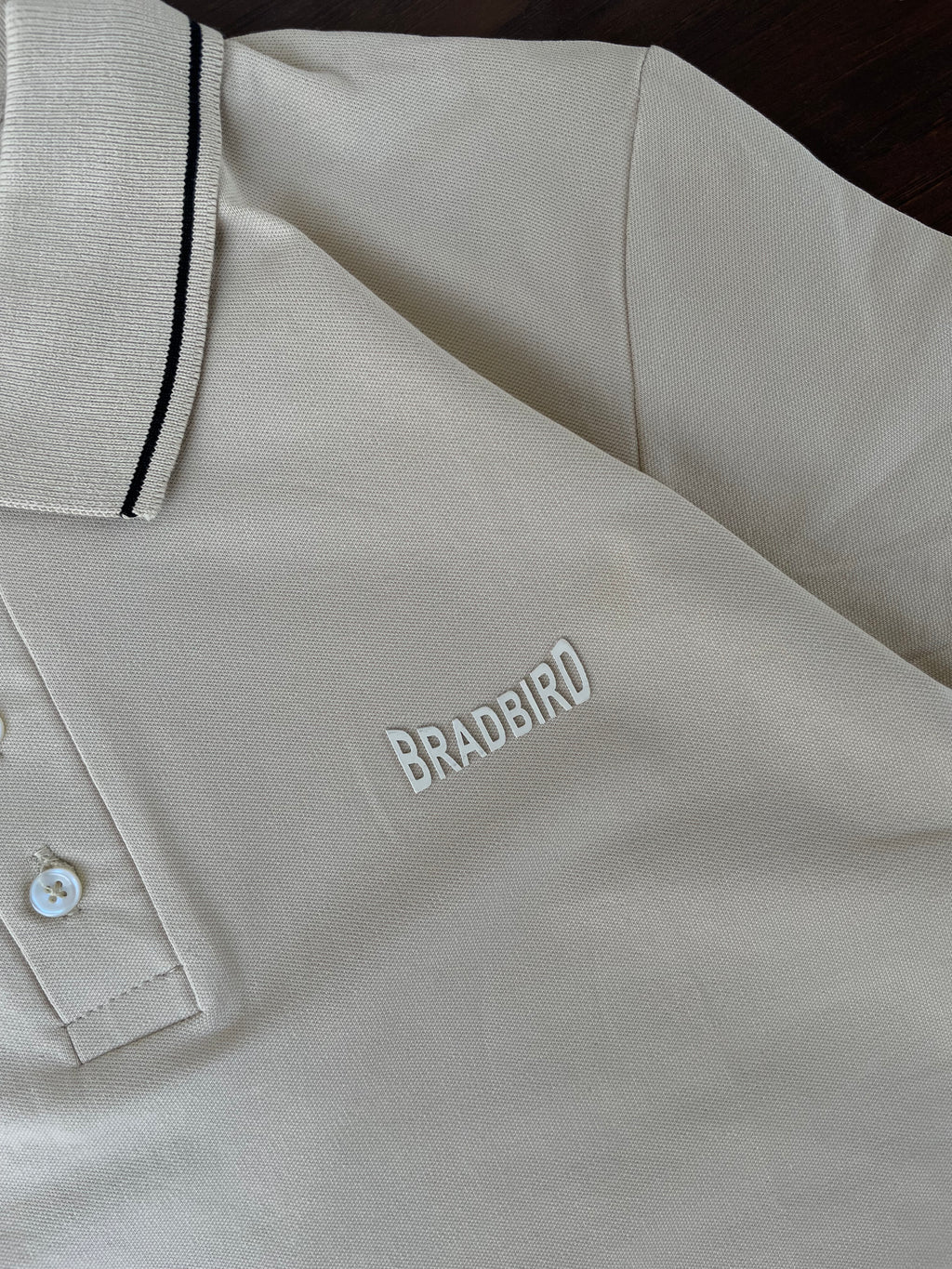 Cream polo shirt close-up of Bradbird chest logo, button placket, navy-trim collar edge.