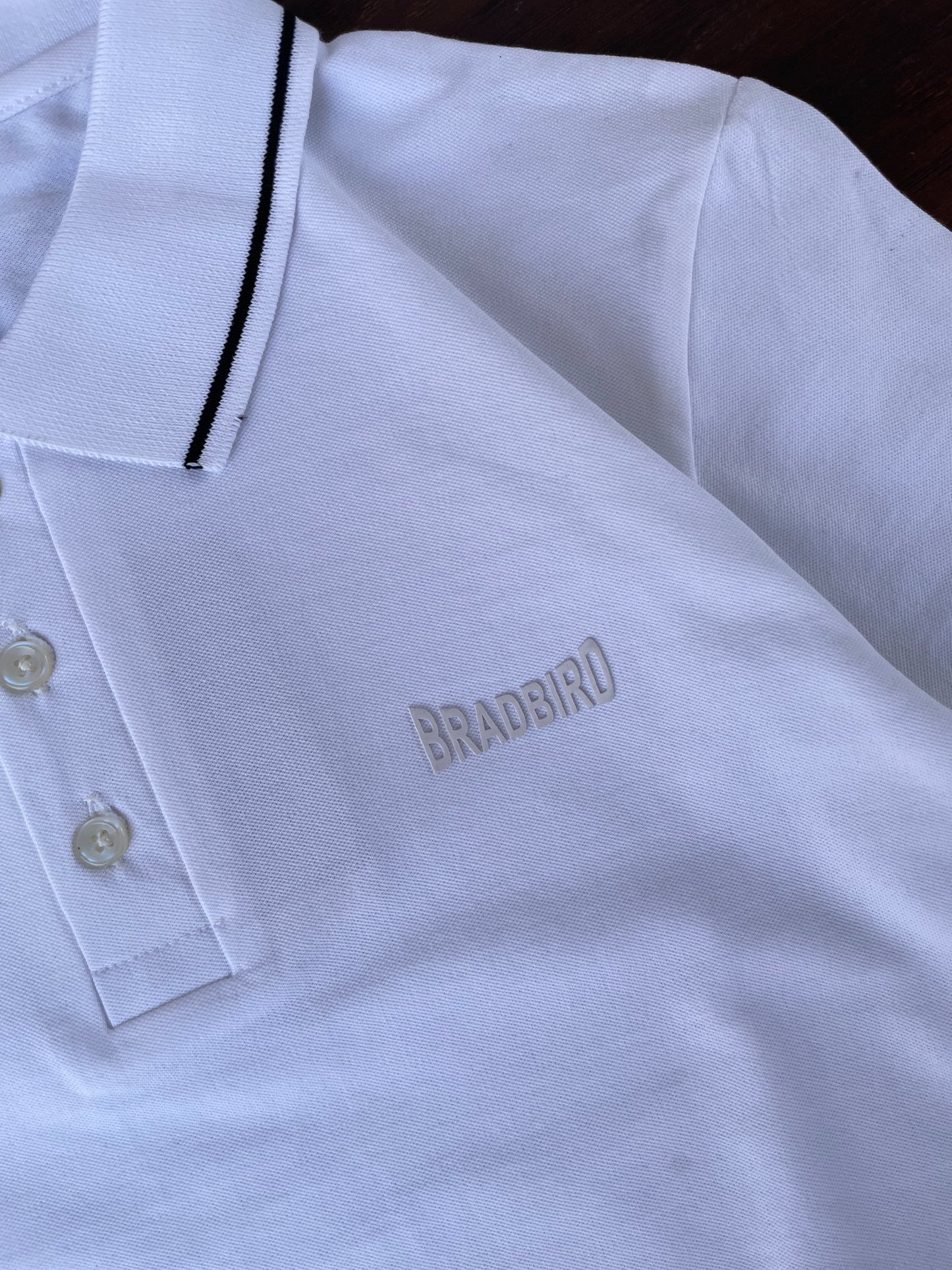 White polo shirt close-up: black-trim placket and small chest emblem, two-button