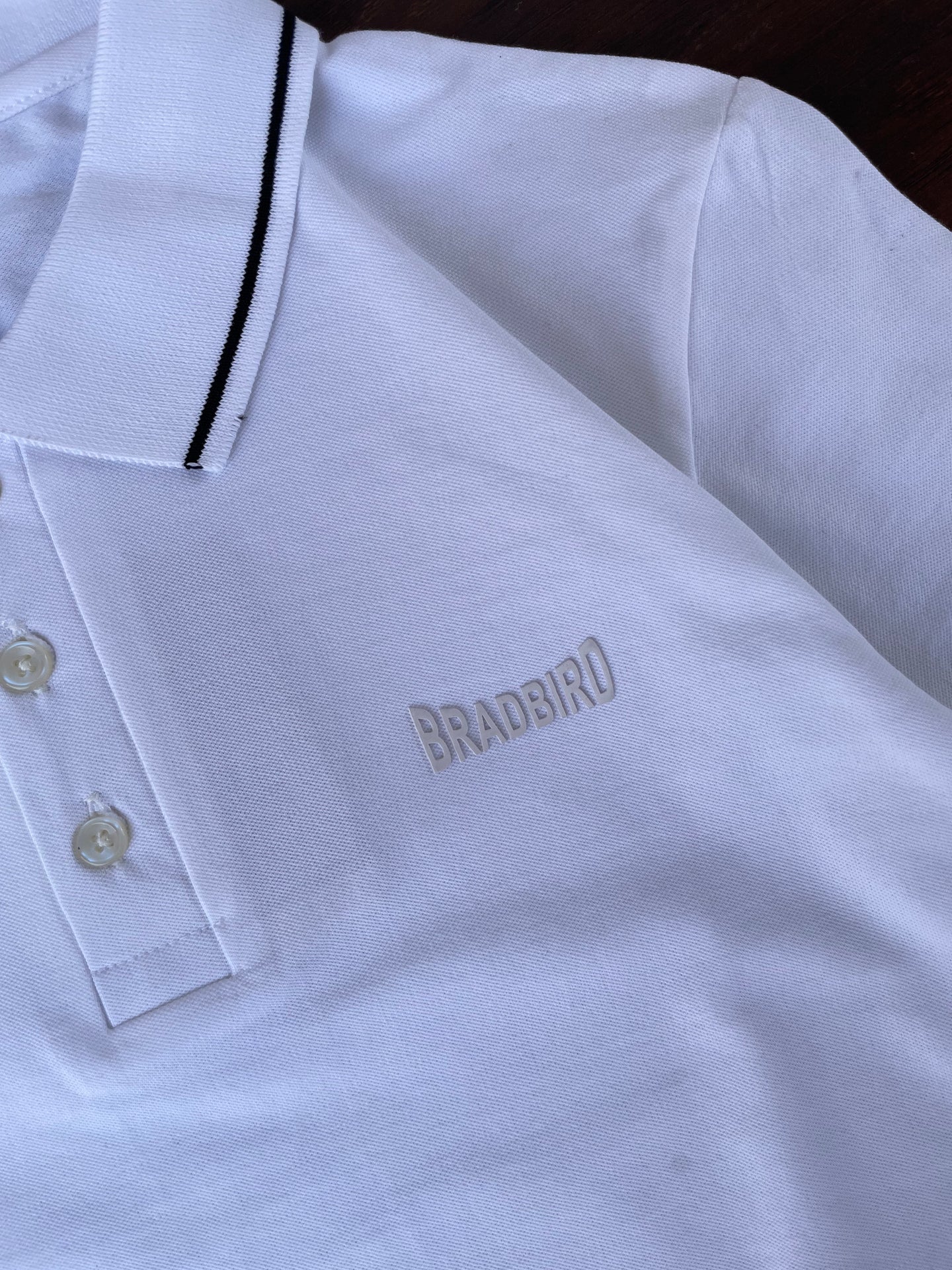 White polo shirt close-up: black-trim placket and small chest emblem, two-button