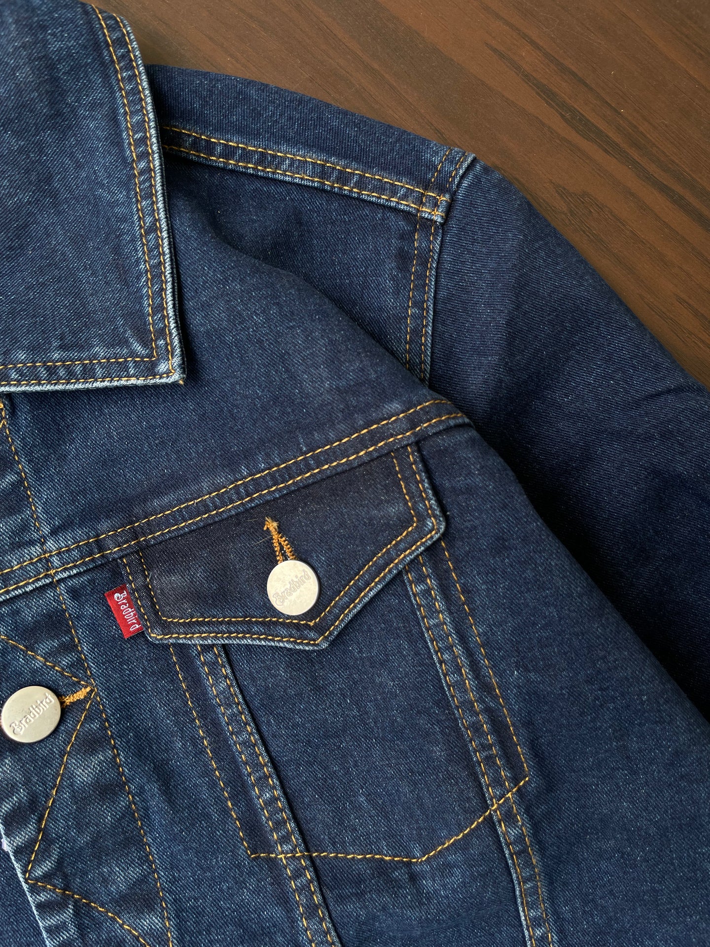 Denim jacket chest pocket detail with metal snap, yellow stitching, and brand patch