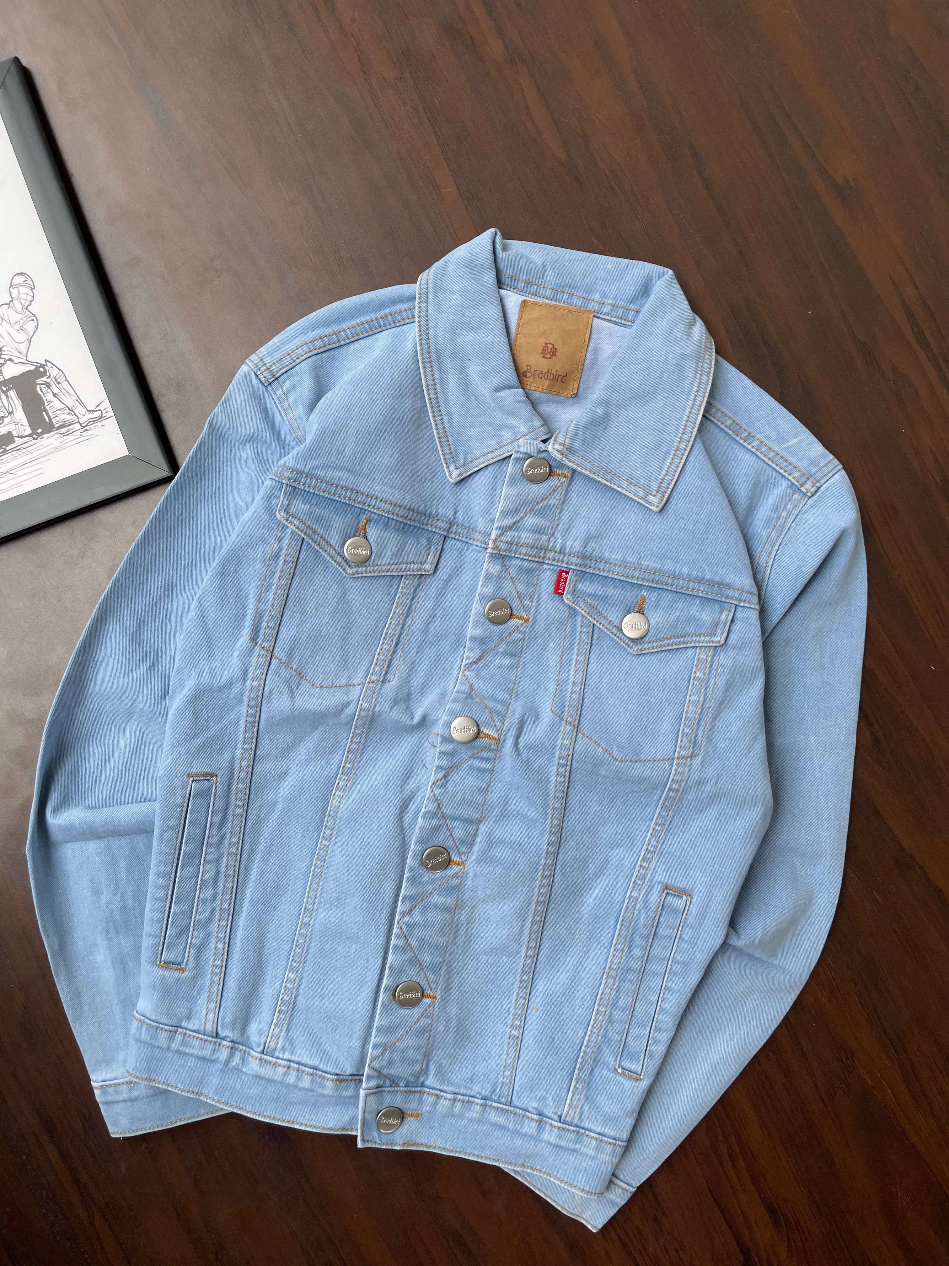 Light-wash denim jacket laid flat, featuring buttoned chest pockets and front placket.