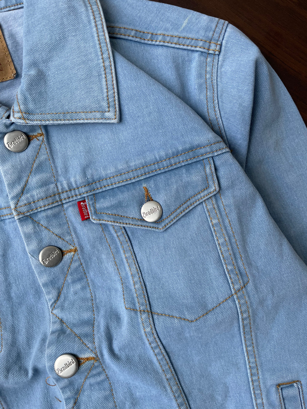 Close-up of light-wash denim jacket pocket with metal buttons and stitched seams.